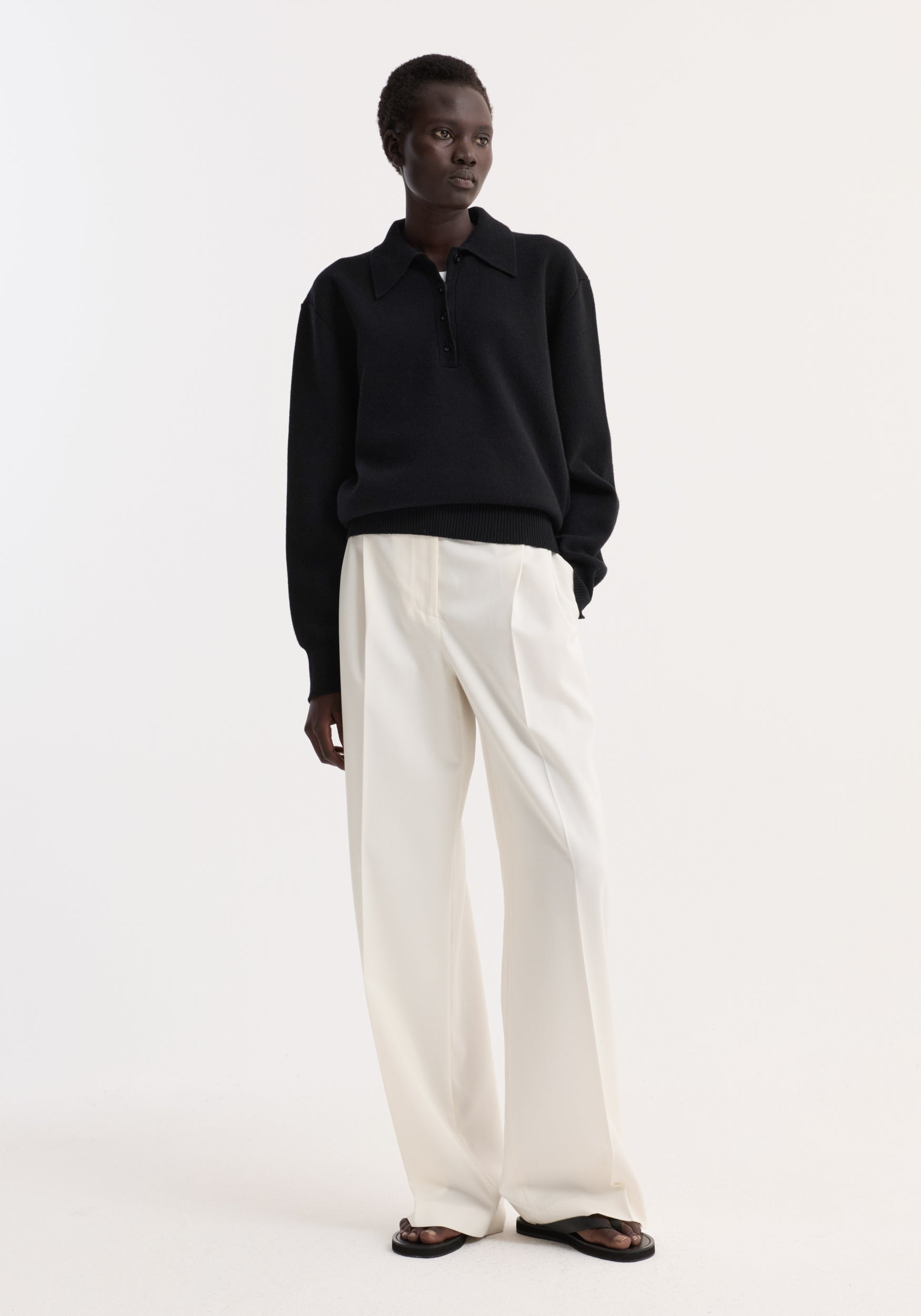 Model wearing a Compact knitted polo | black with collared, three-button front, and relaxed fit, paired with wide-leg trousers and sandals against a neutral background.