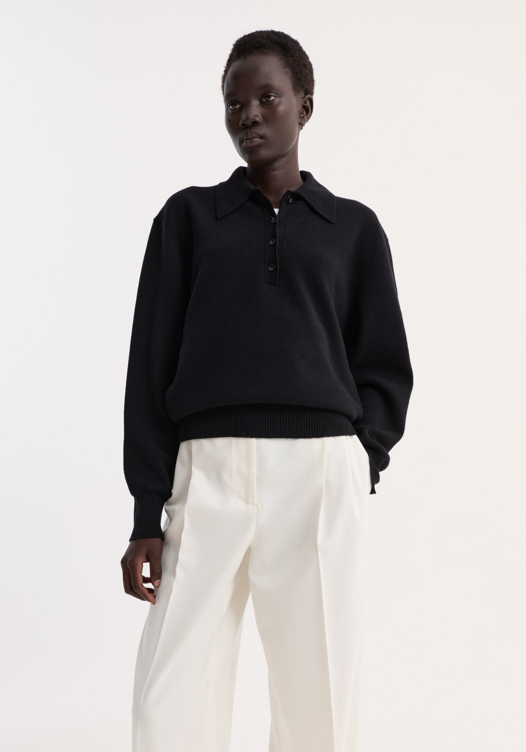 Model wears the Compact knitted polo in a wool-cashmere blend, featuring a structured collar, three-button closure, and rounded drop shoulder, paired with wide-leg trousers.