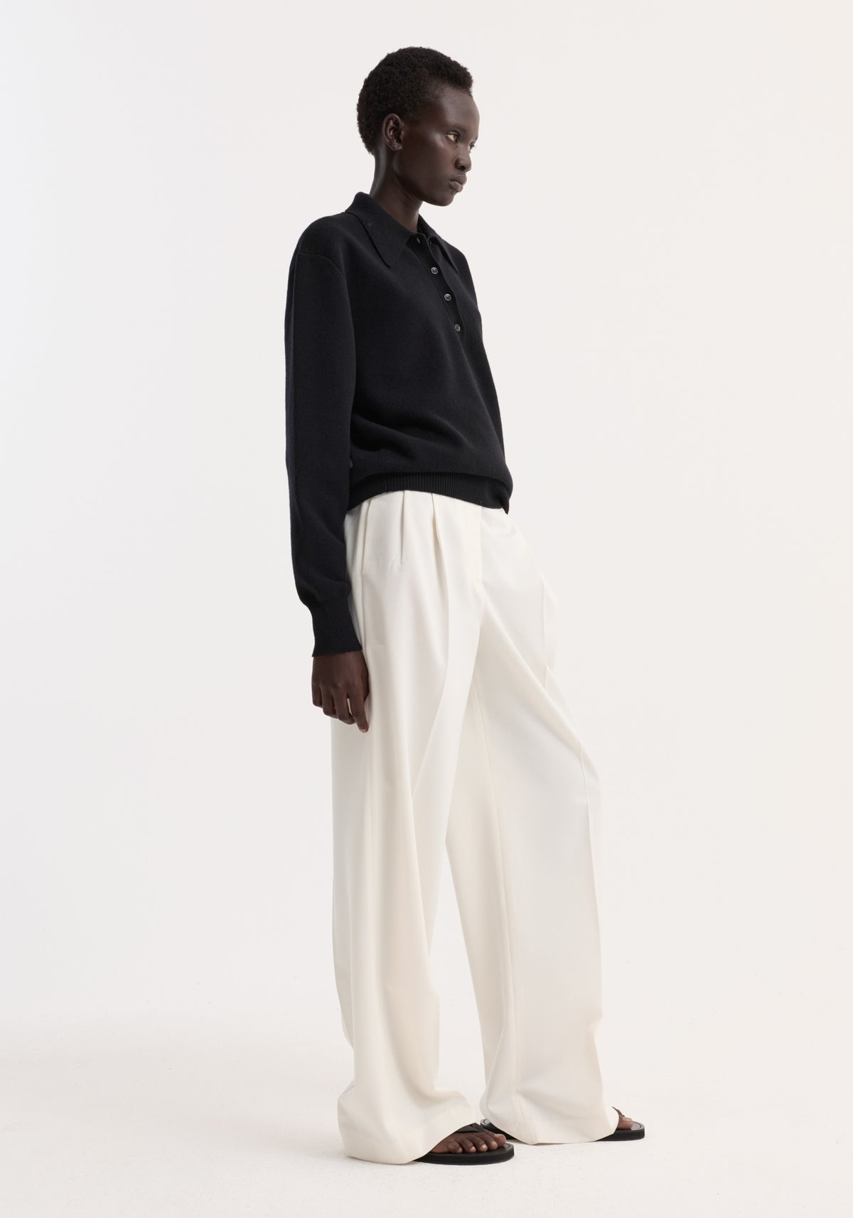 Model in profile wearing the Compact knitted polo in black with a button-up collar, paired with wide-leg trousers and black toe-loop sandals.