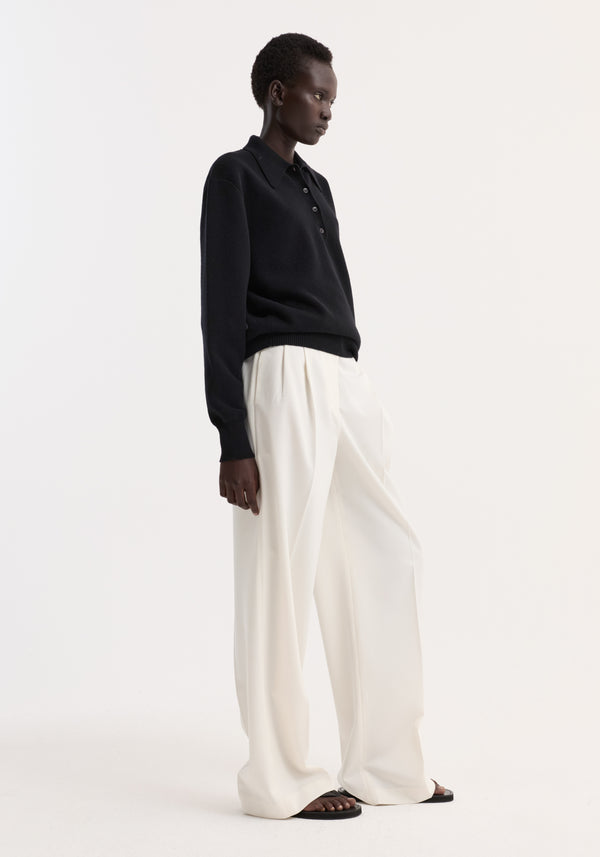 Model in profile wearing the Compact knitted polo in black with a button-up collar, paired with wide-leg trousers and black toe-loop sandals.