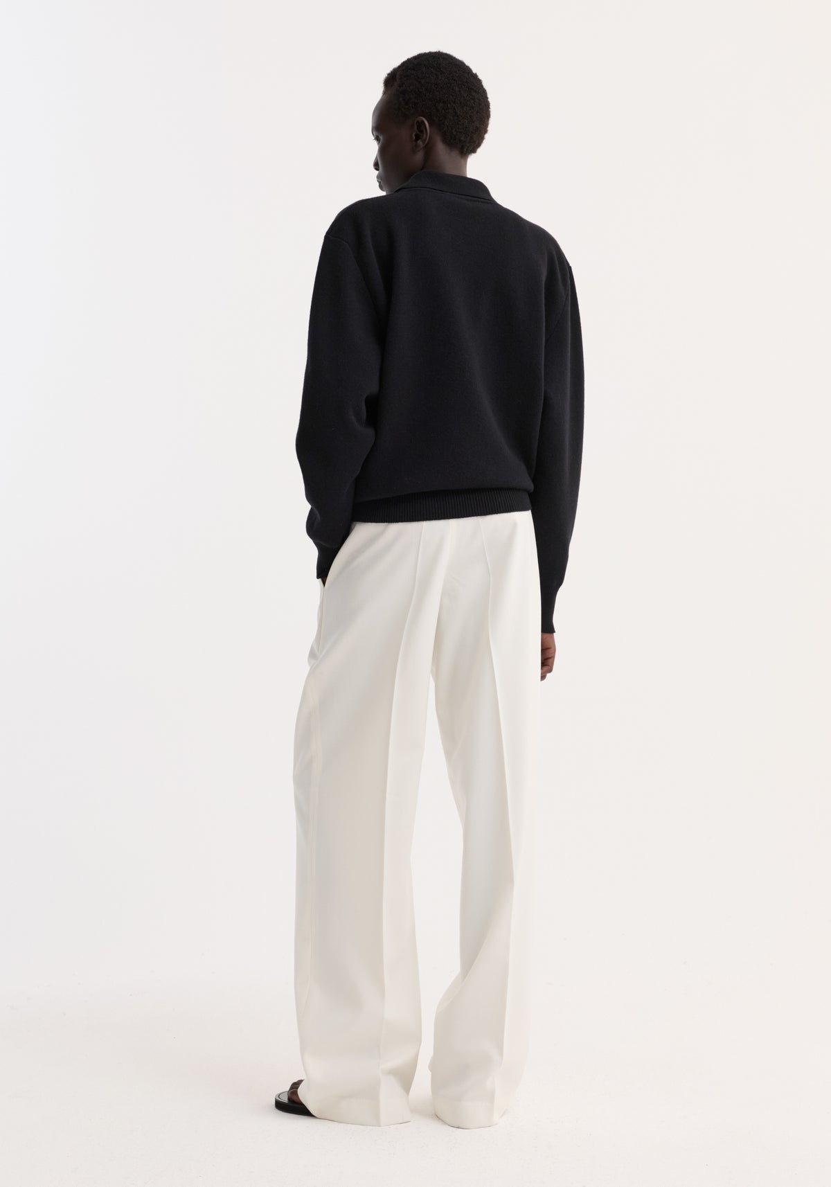 Model wearing Compact knitted polo with rounded drop shoulders, structured collar, and three-button front closure, paired with wide-leg trousers, viewed from behind.