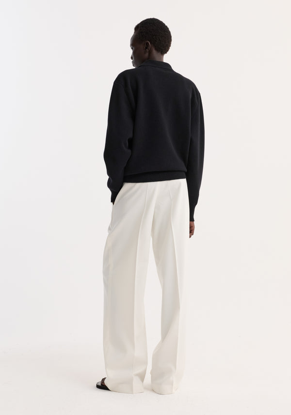 Model wearing Compact knitted polo with rounded drop shoulders, structured collar, and three-button front closure, paired with wide-leg trousers, viewed from behind.