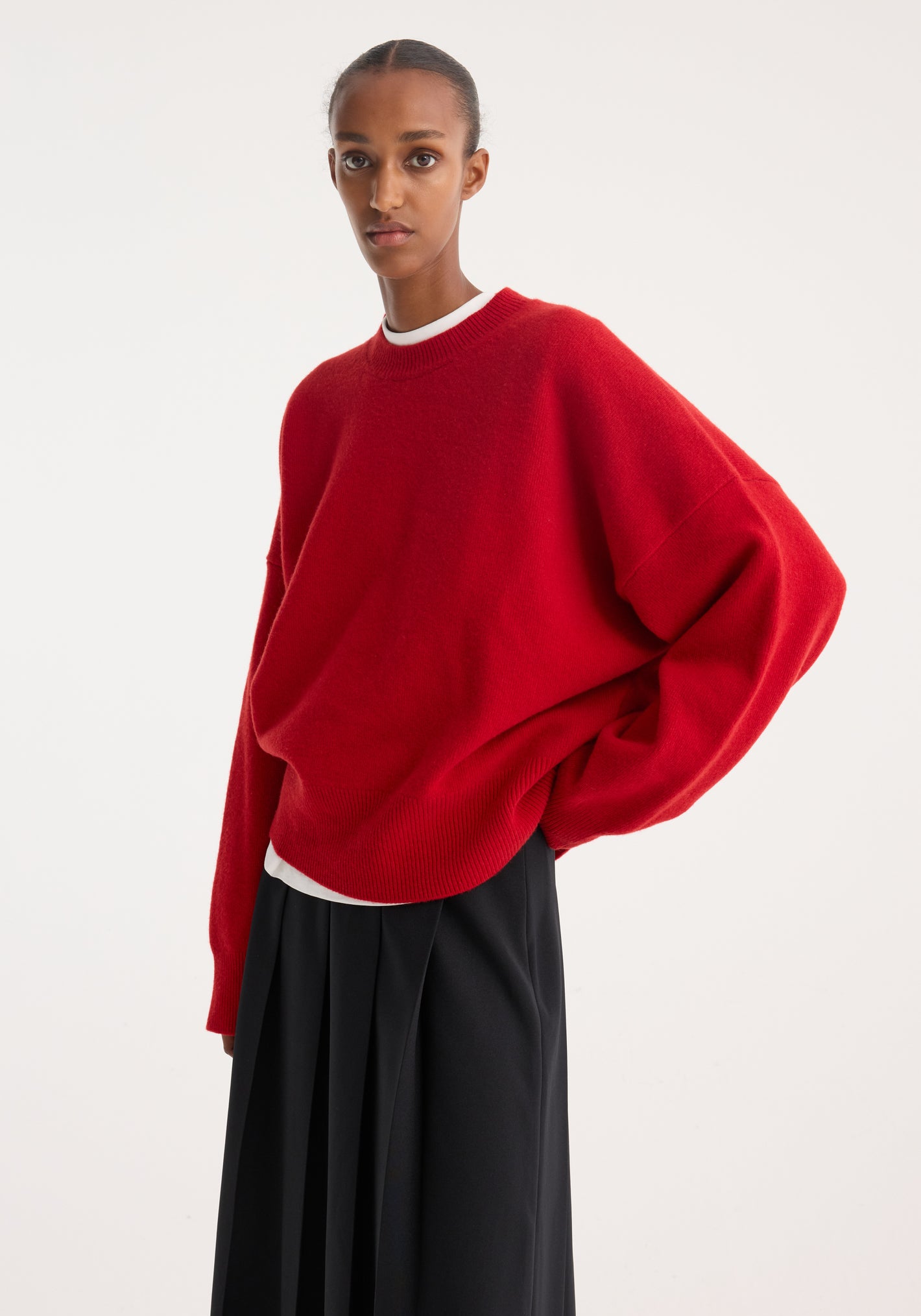 Boiled wool crewneck | rosso: A minimalist portrait shows the relaxed fit of the oversized red crewneck sweater, highlighting its drop shoulder design over a pleated skirt.