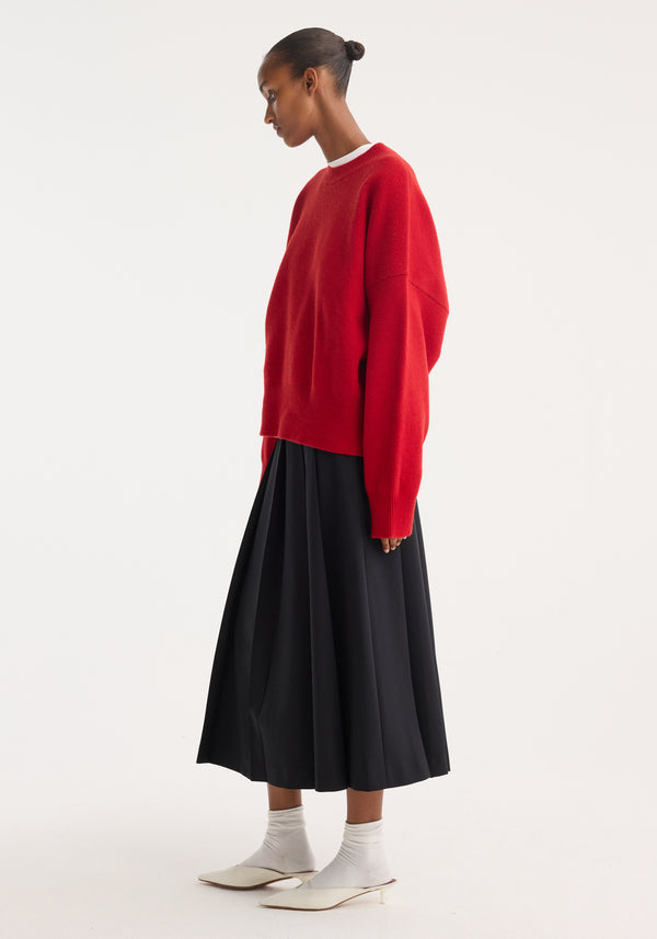 Model in an oversized Boiled wool crewneck | rosso with a relaxed fit, paired with a black pleated midi skirt, white socks, and mule shoes.