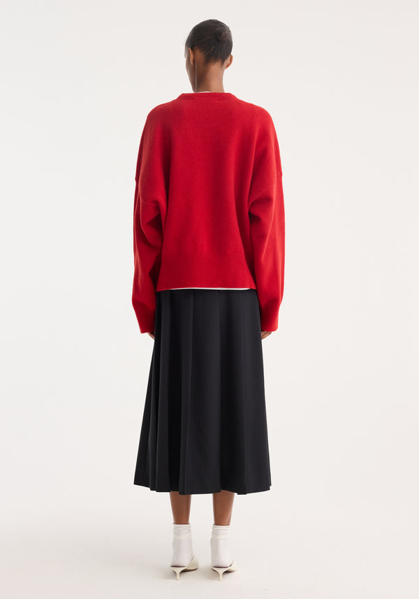 The person wears a Boiled wool crewneck | rosso, showcasing its relaxed fit and drop shoulder, paired with a black pleated midi skirt and white shoes.