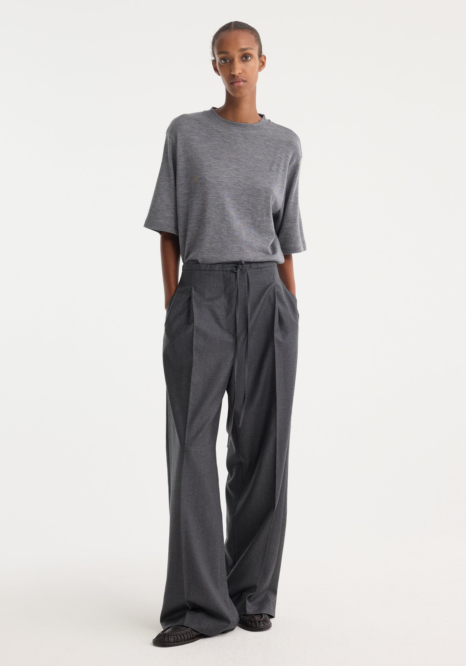 Tall person wearing Relaxed soft wool trousers, dark grey melange, with wide legs, deep pleats, and drawstring waist, showcasing a comfortable, elegant fit.
