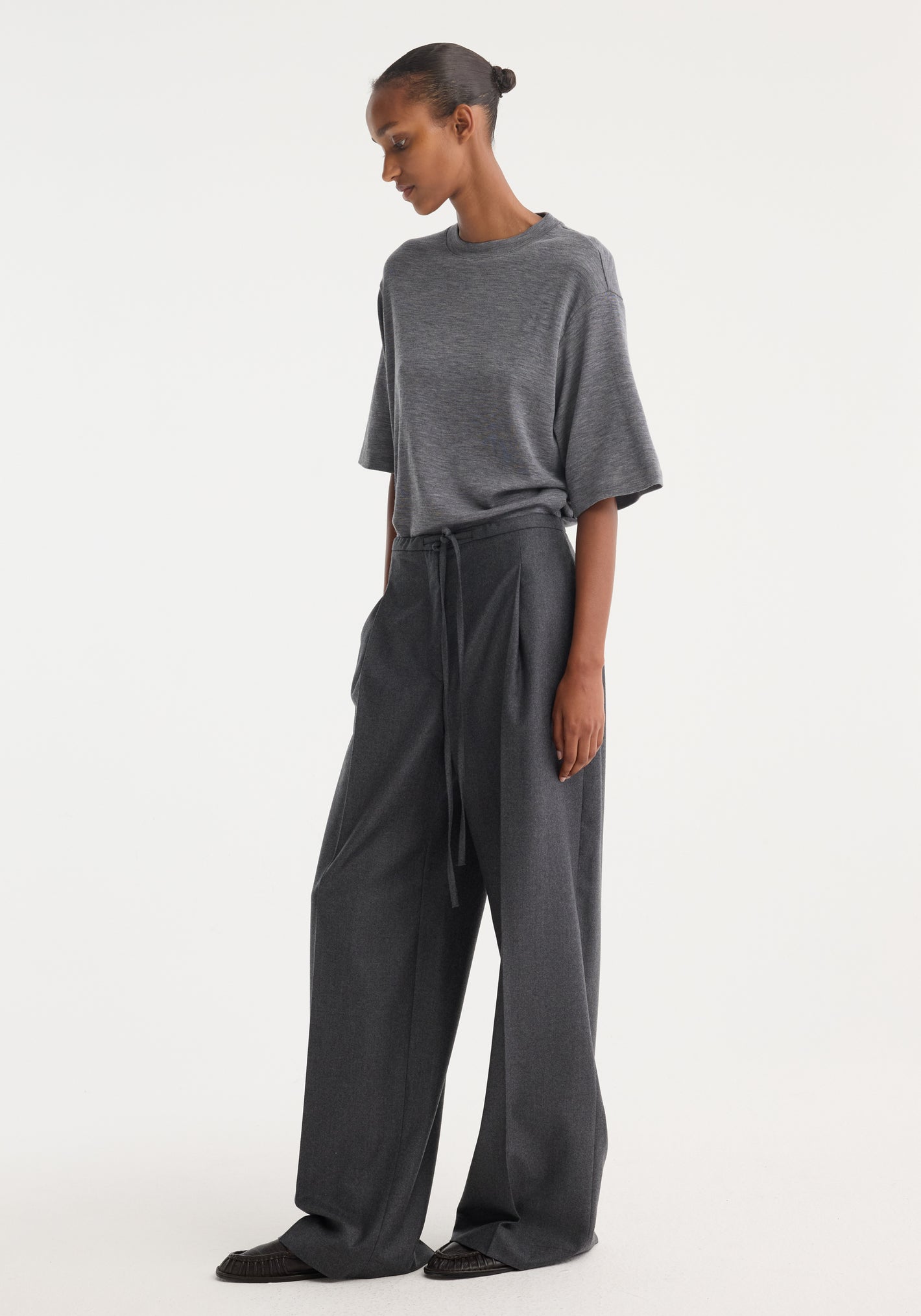 Model wearing relaxed soft wool trousers with a wide-leg silhouette, drawstring waistband, and side pockets, styled with a charcoal-gray T-shirt and slip-on shoes.