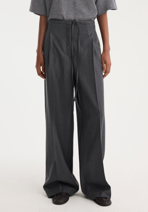 Relaxed soft wool trousers in dark grey melange, featuring a wide-leg design and drawstring waistband, paired with a tucked-in heather-gray top and moccasin-style shoes.