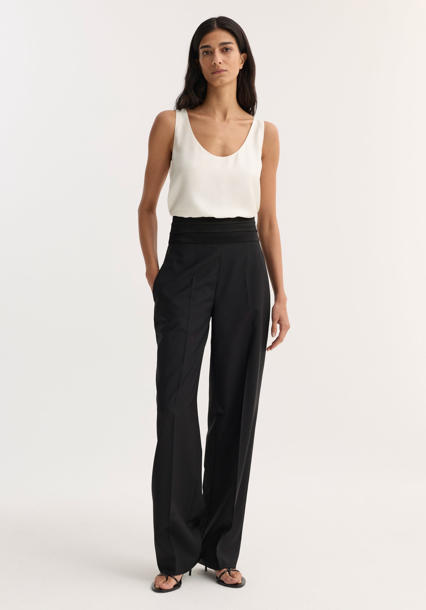Model wearing Cummerbund trousers in black, featuring a structured satin waistband and shell-covered buttons, paired with a sleeveless scoop-neck top and heeled sandals.