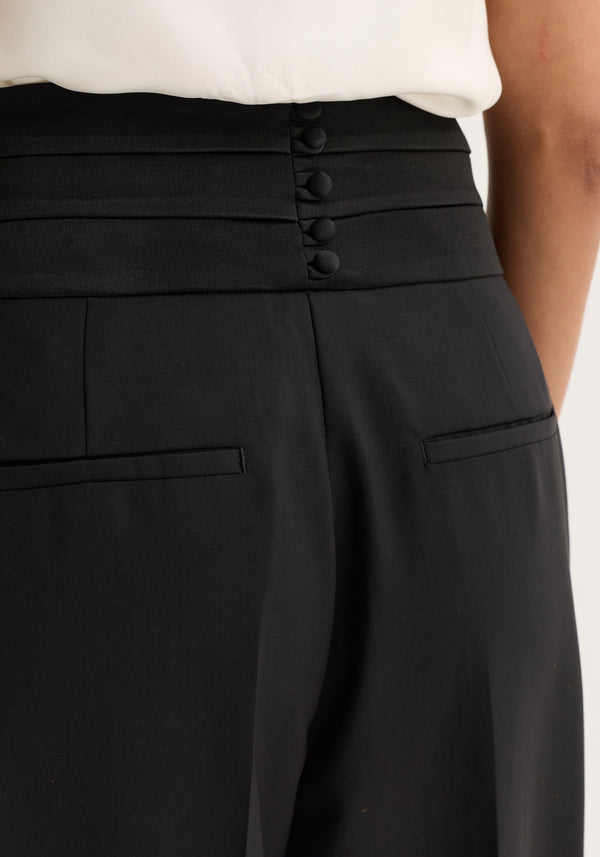 Close-up of Cummerband trousers in black, showcasing a wide, pleated satin waistband with shell-covered buttons at the back and slit pockets below.