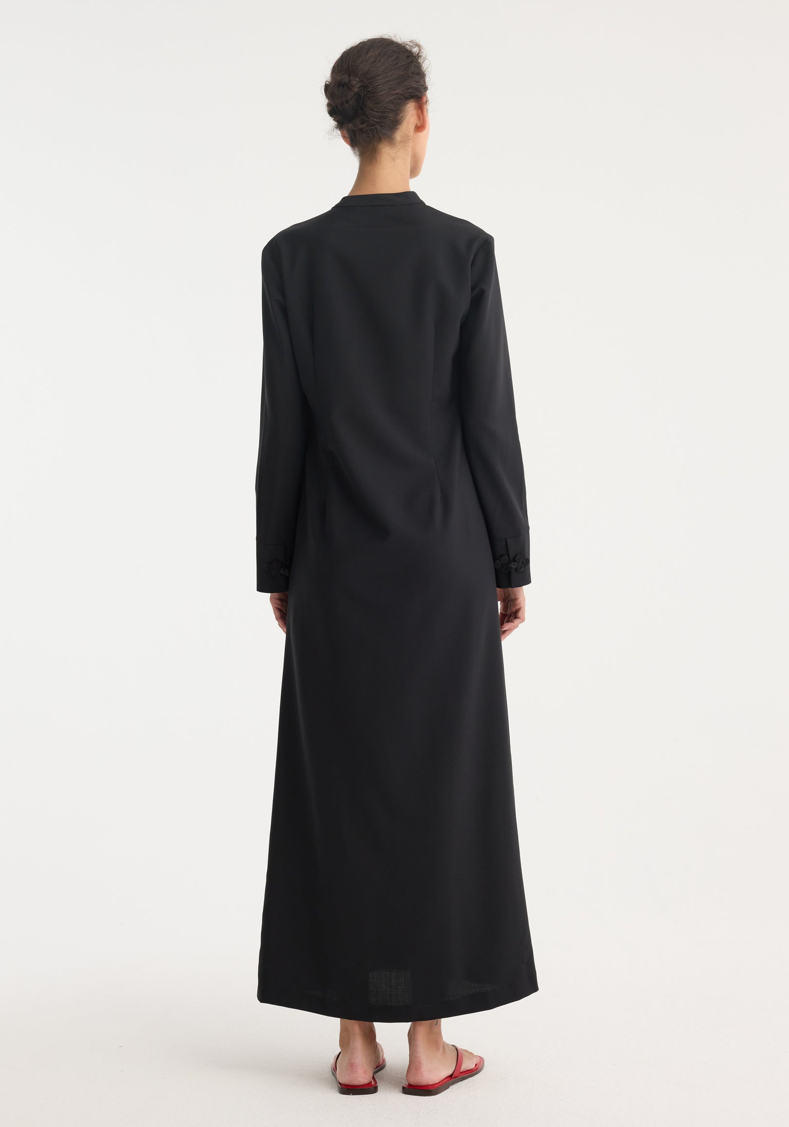 Pankou closure dress | black: Back view of a woman in a floor-length, long-sleeve dress with asymmetrical pankou closures and side split, hair in bun, red sandals.