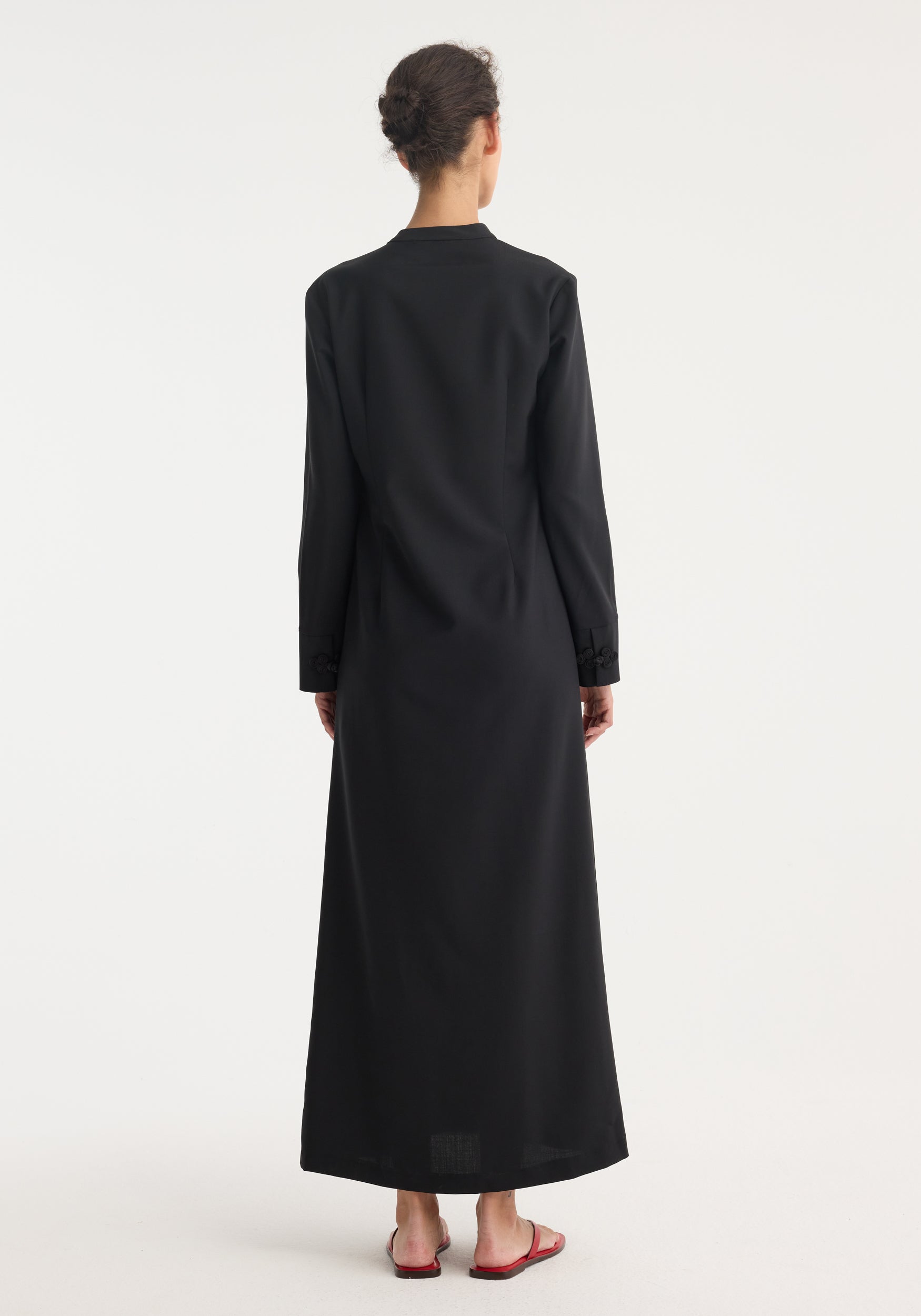 Pankou closure dress | black: Back view of a woman in a floor-length, long-sleeve dress with asymmetrical pankou closures and side split, hair in bun, red sandals.