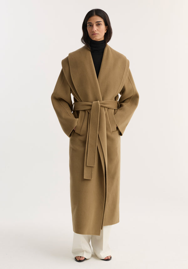 Model wears Signature double-faced scarf coat in camel, featuring a double-layered scarf collar, double-breasted design, side pockets, over black turtleneck and white trousers.
