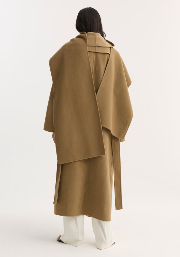 Person wearing the Signature double-faced scarf coat in camel, showcasing its double-layered scarf collar and long, tailored silhouette, paired with wide-leg trousers and sandals.