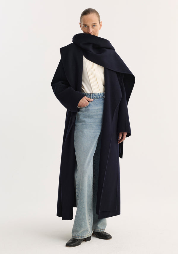 Signature double-faced scarf coat in navy with a striking double-layered scarf collar, worn by a model with straight-leg jeans and black ankle boots.