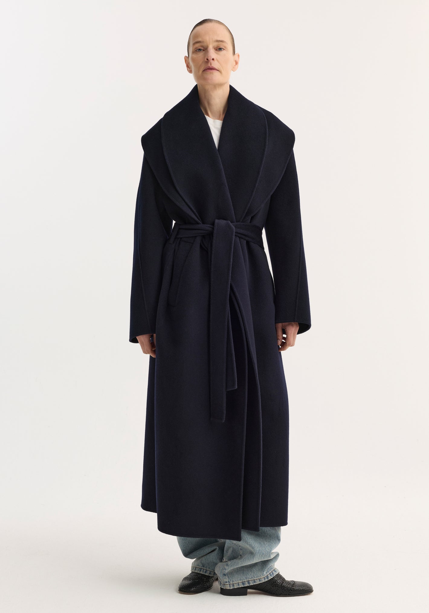 Model wears Signature double-faced scarf coat in navy, showcasing an oversized shawl collar, self-tie belt, and long silhouette over light-wash jeans and black shoes.