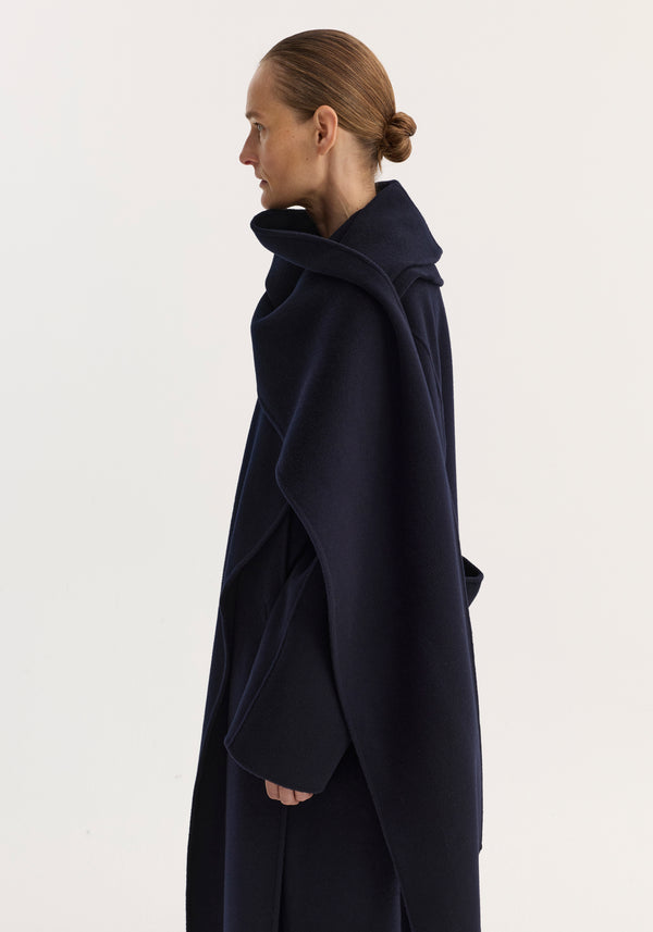 Woman in profile wearing the Signature double-faced scarf coat in navy, featuring a double-layered scarf collar, oversized sleeves, and voluminous lapels against an off-white background.