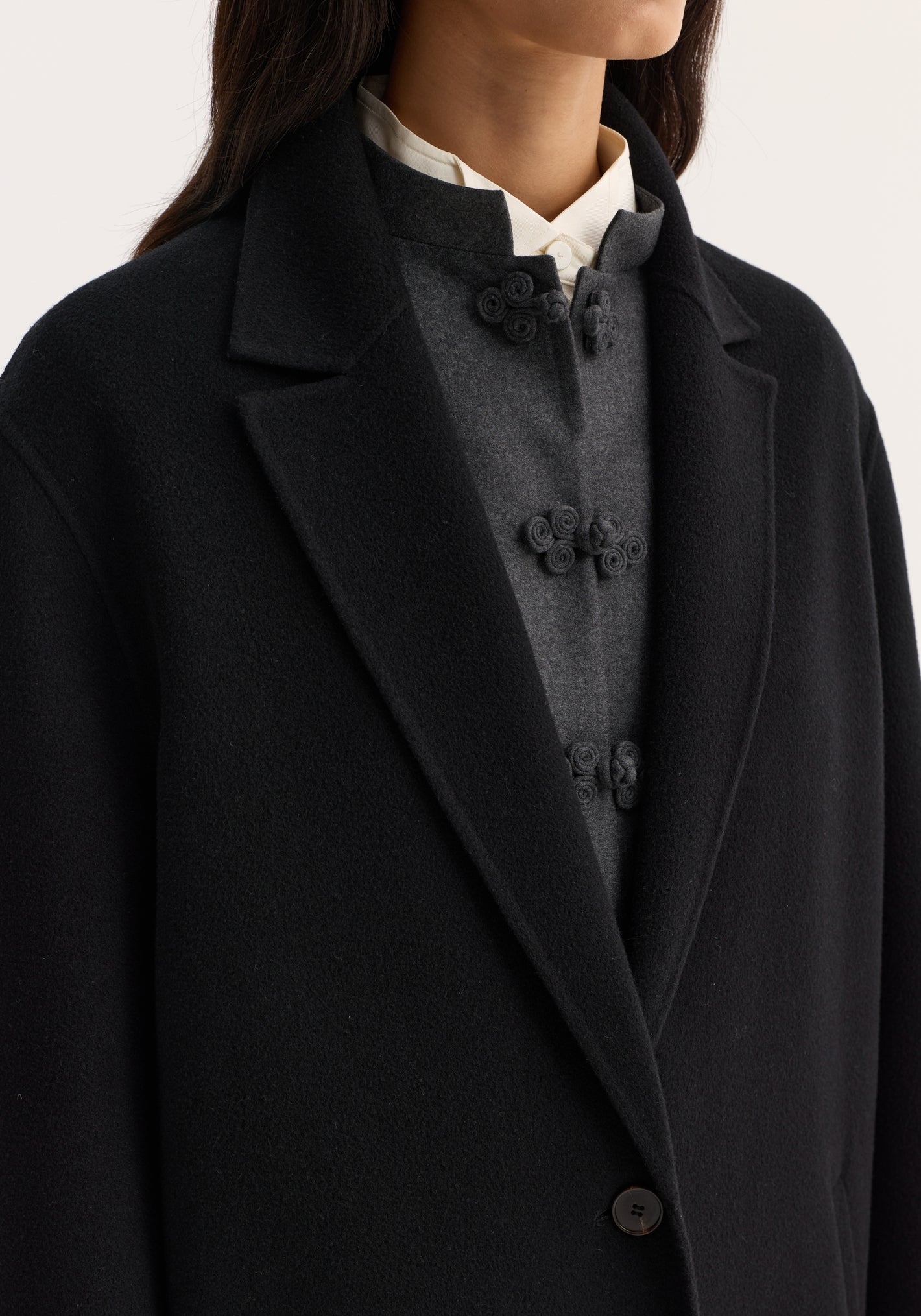 Double-faced coat in black with a single button, notched lapel, layered over a grey Mandarin-collar top and white collared shirt.