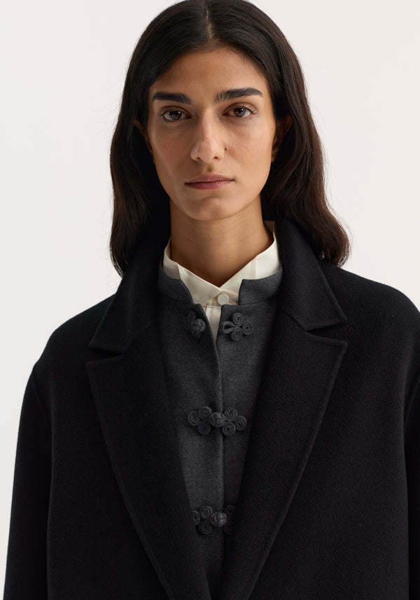 Double-faced coat in black, single-breasted with frog-style buttons, worn by a model. Made from 100% wool, featuring a straight silhouette and side pockets.