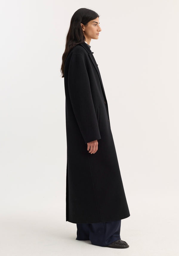Side-profile of a person in a Double-faced coat | black, showcasing its full-length, single-breasted design, and discreet side pockets, paired with wide-leg trousers and slip-on shoes.