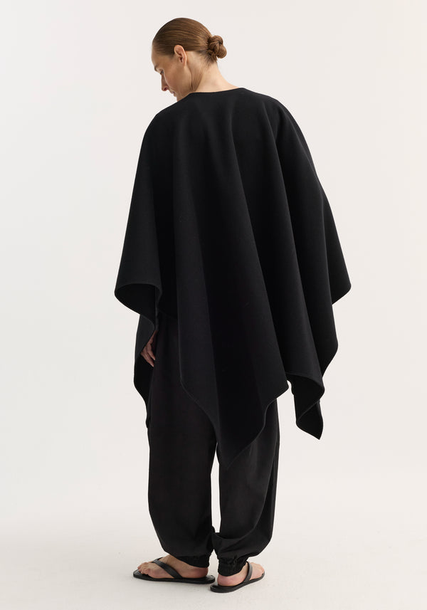 Model wearing Draped scarf poncho in black, showcasing its loose wool design and flowing silhouette, paired with black relaxed-fit pants and flip-flop sandals.