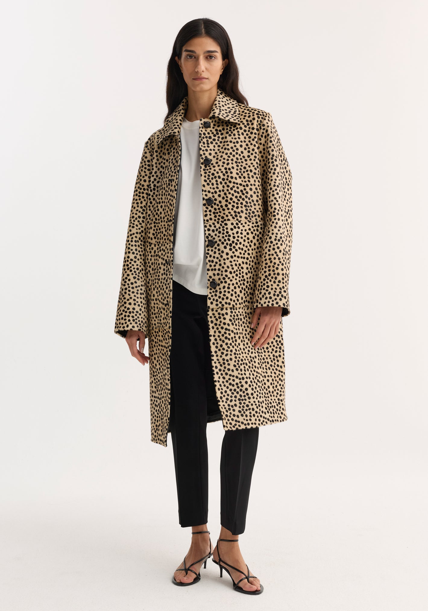 Model in a cheetah-print pony hair coat with leather-knotted buttons, white top, black trousers, and strappy sandals. The coat features hidden pockets and a structured silhouette.