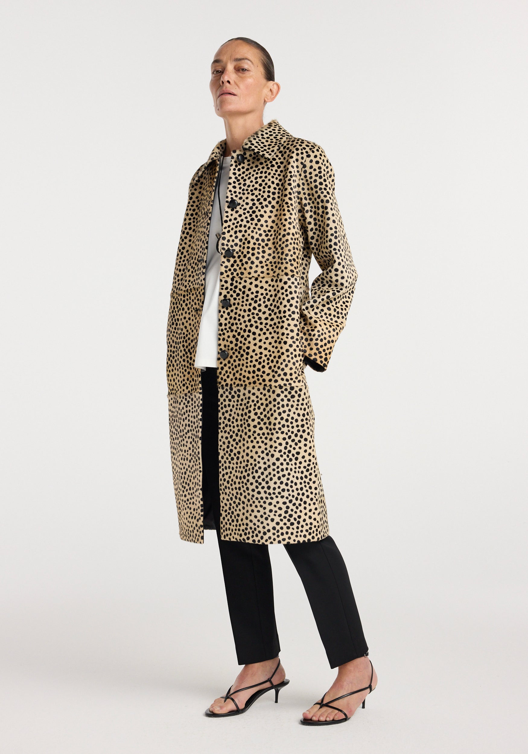 Woman wearing a long pony hair coat | cheetah with black spots on a tan background, paired with black pants, white top, and black strappy heels, standing against a white backdrop.