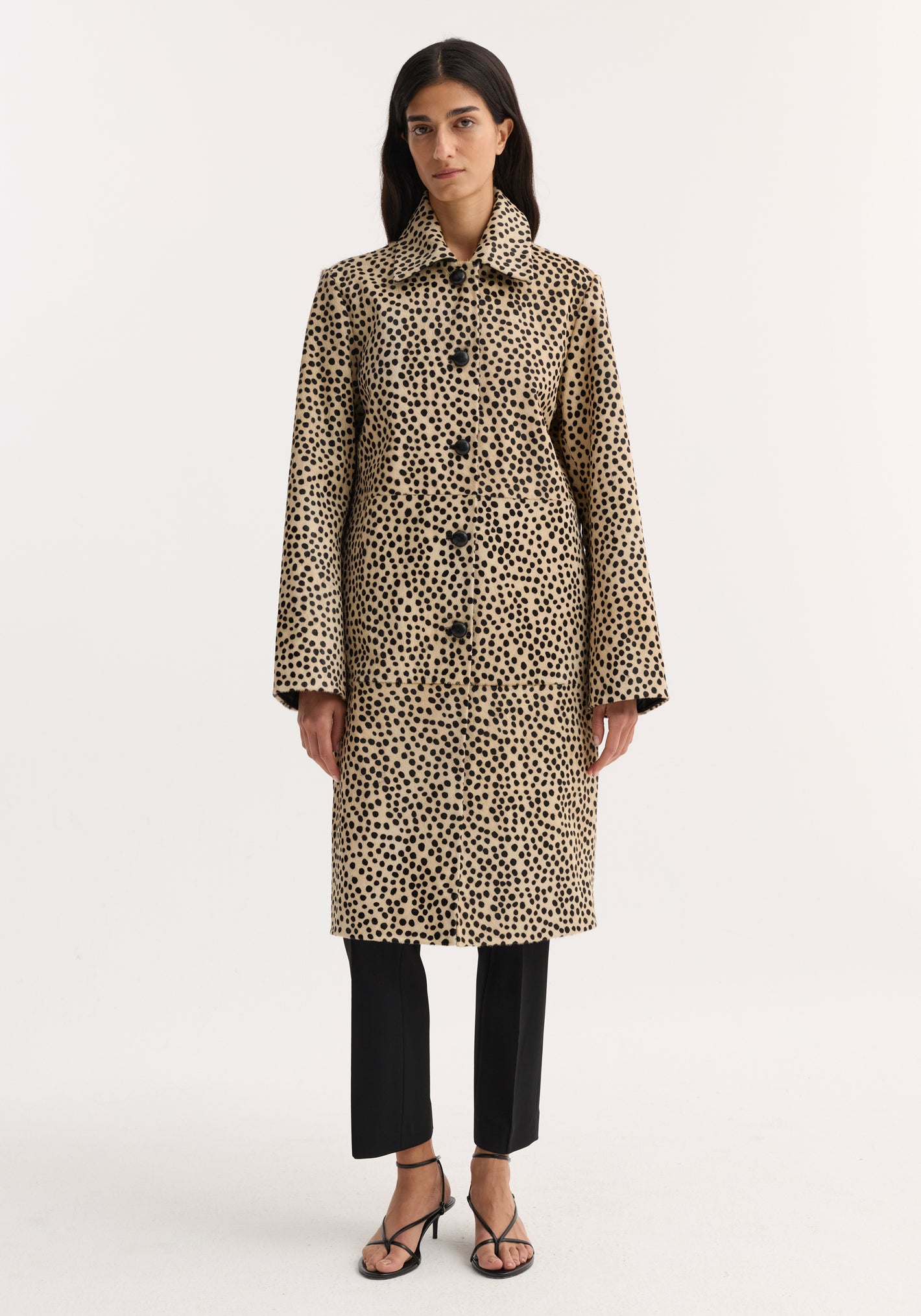 Model wearing a Pony hair coat | cheetah, featuring a knee-length cheetah-print design with leather-knotted buttons, over black trousers and sandals. Structured, refined silhouette.