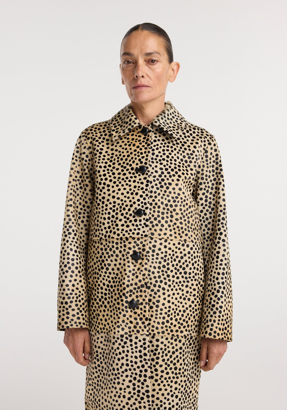 Woman wearing a long Pony hair coat | cheetah with black spotted pattern, buttoned front, and classic collar, standing against a plain white background.