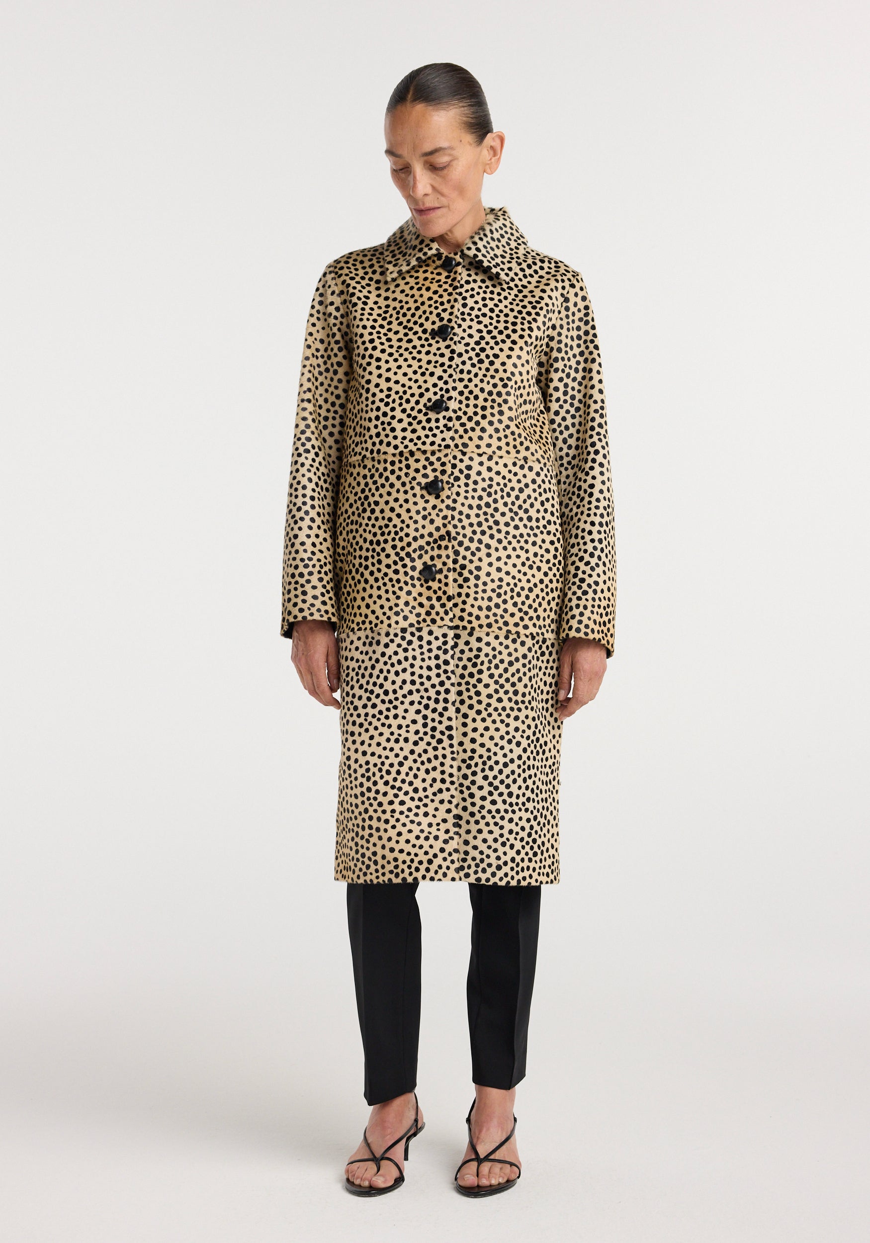 Woman wearing a knee-length Pony hair coat | cheetah in tan with black spots, buttoned front, long sleeves, and a collar, styled with black pants and strappy heels.