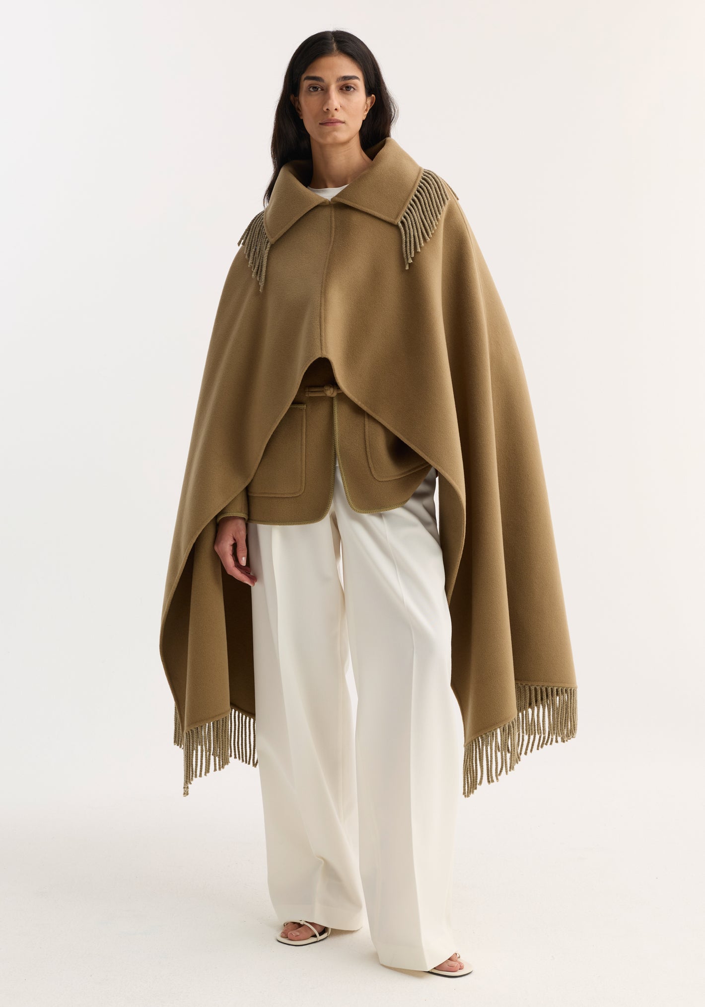 Woman wearing Fringe scarf poncho in camel, featuring fringe trim and a loose silhouette, over a belted vest, paired with white trousers and sandals.