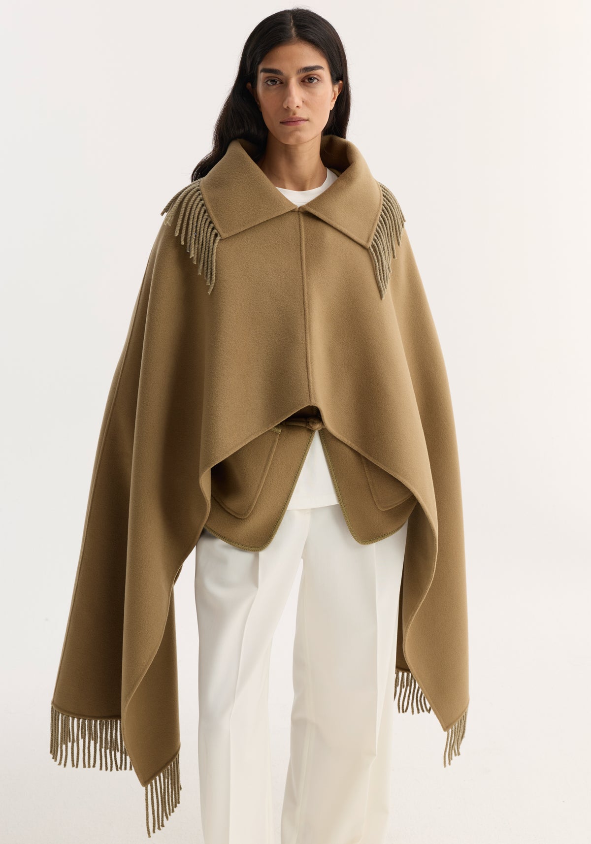 Model wearing Fringe scarf poncho in camel, showcasing its double-faced wool–cashmere blend, wide collar, and fringed trim over a minimalist white ensemble.