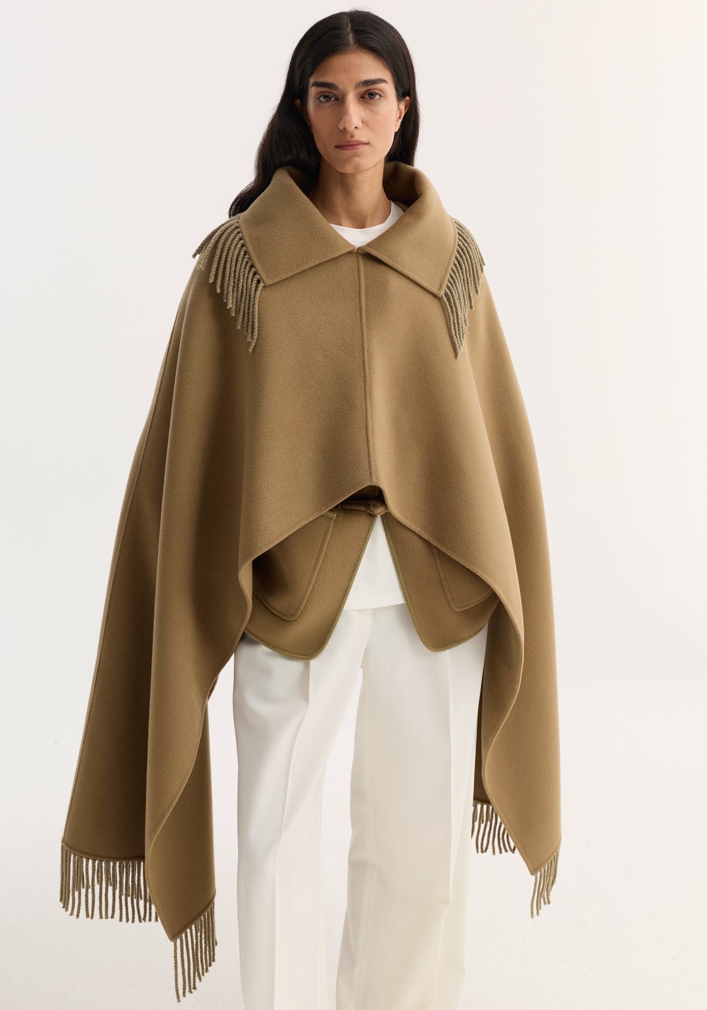Model wearing Fringe scarf poncho in camel, showcasing its double-faced wool–cashmere blend, wide collar, and fringed trim over a minimalist white ensemble.