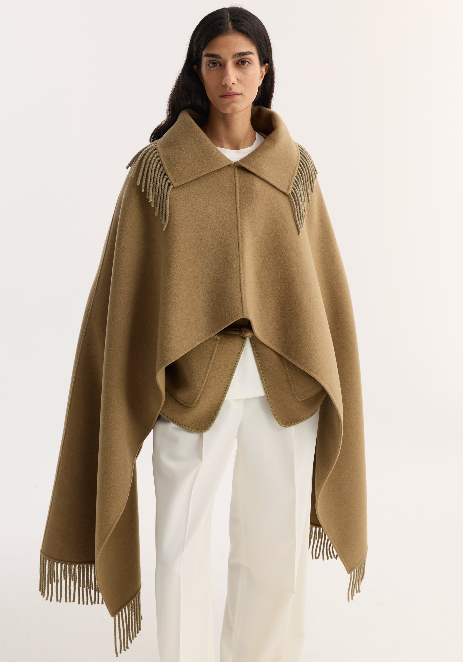 Model wearing Fringe scarf poncho in camel, showcasing its double-faced wool–cashmere blend, wide collar, and fringed trim over a minimalist white ensemble.