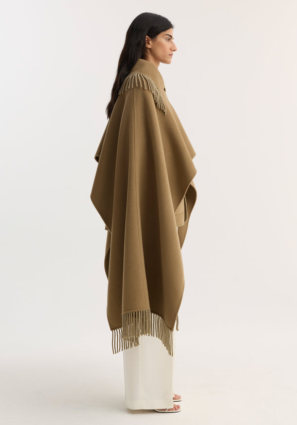 Model wearing Fringe scarf poncho in camel, a wool-cashmere blend with long fringe trim, paired with off-white trousers and white sandals, showcasing its elegant drape.