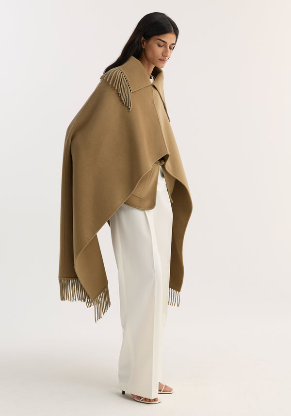 Model in a Fringe scarf poncho | camel, showcasing a wool-cashmere blend with fringe detailing, paired with white trousers and sandals, exuding an elegant, editorial vibe.