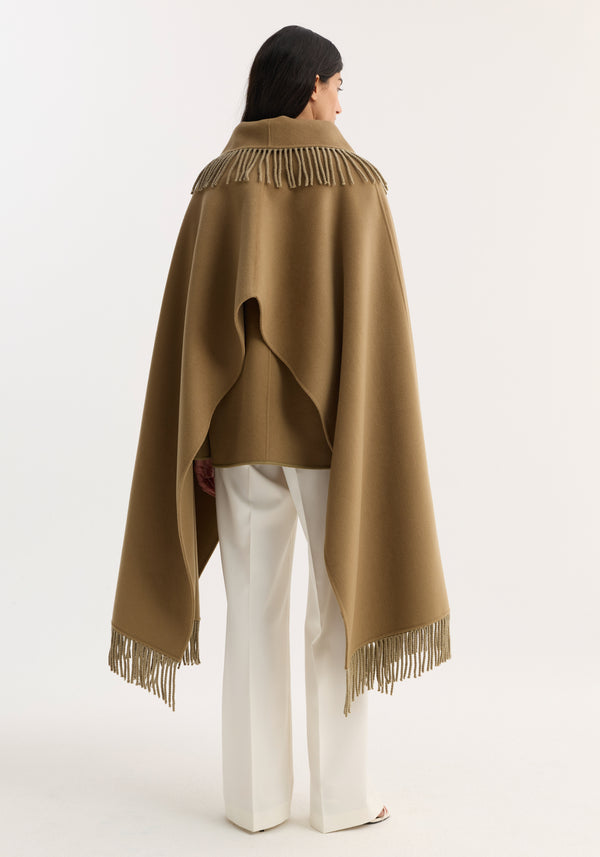 Model wearing Fringe scarf poncho in camel, featuring a wool-cashmere blend with long fringe detailing. Styled with white trousers, offering an elegant, warm silhouette.