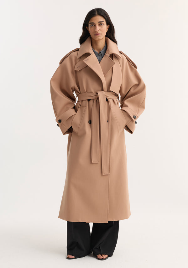 Woman wearing an oversized classic trench with structured detailing, wide collar, epaulettes, and cinched belt; black trousers peek from beneath, completing the modern look.