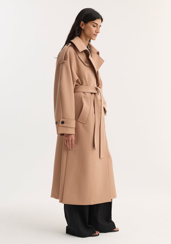 Model wearing Oversized classic trench | powder, showcasing its belted waist, wide lapels, and epaulet-style shoulder tabs, paired with black trousers and open-toed sandals.
