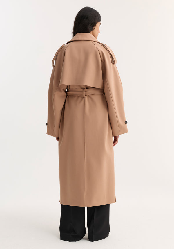 Back view of a model in an Oversized classic trench | powder, showcasing the belted, camel trench coat with storm flap and epaulettes, paired with black wide-leg pants.