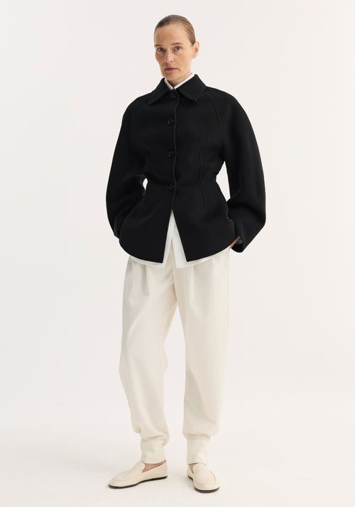 Woman wearing a double-faced sculptural jacket with button closures and sculpted silhouette, paired with ankle-gathered trousers and loafers, hair pulled back neatly.