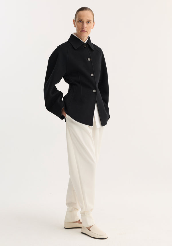 Light-skinned adult wears a Double-faced sculptural jacket in black, featuring a cinched waist and oversized sleeves, paired with high-waisted white trousers and cream shoes.
