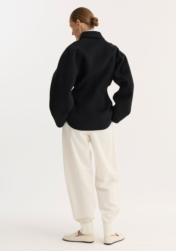 Model in double-faced sculptural jacket with voluminous sleeves, seaming, and button closures, paired with loose joggers and slip-ons, showcasing a tailored, architectural design.