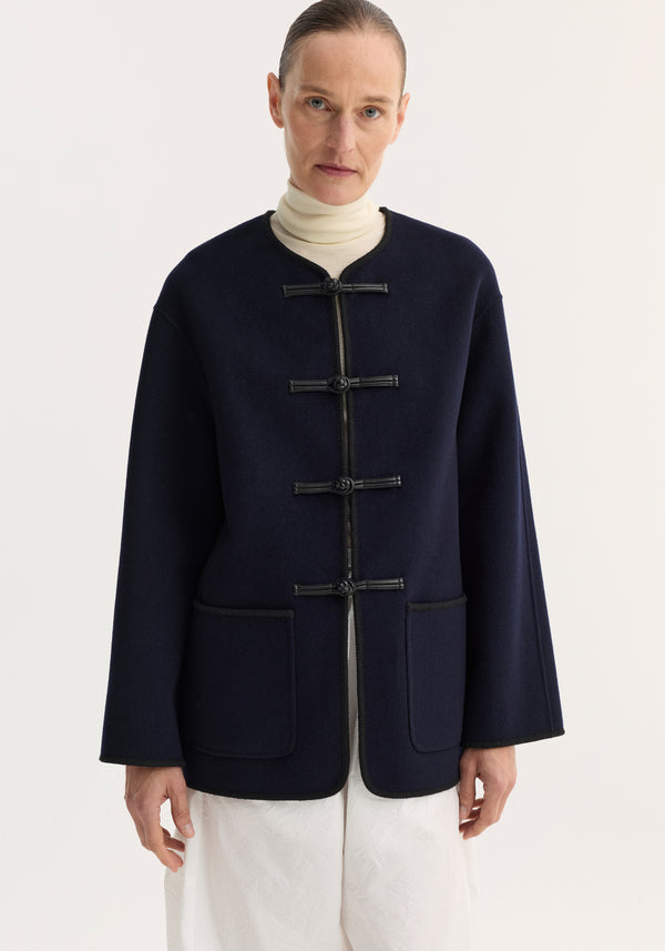 Model in a navy Mandarin double-faced wool jacket with pankou closure, embroidered edges, and patch pockets, paired with a light turtleneck and white pants.