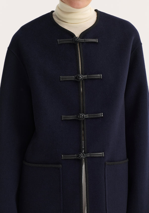 Mandarin double-faced wool jacket in navy with pankou closures, embroidered edges, patch pockets. Model wears it over a light turtleneck.