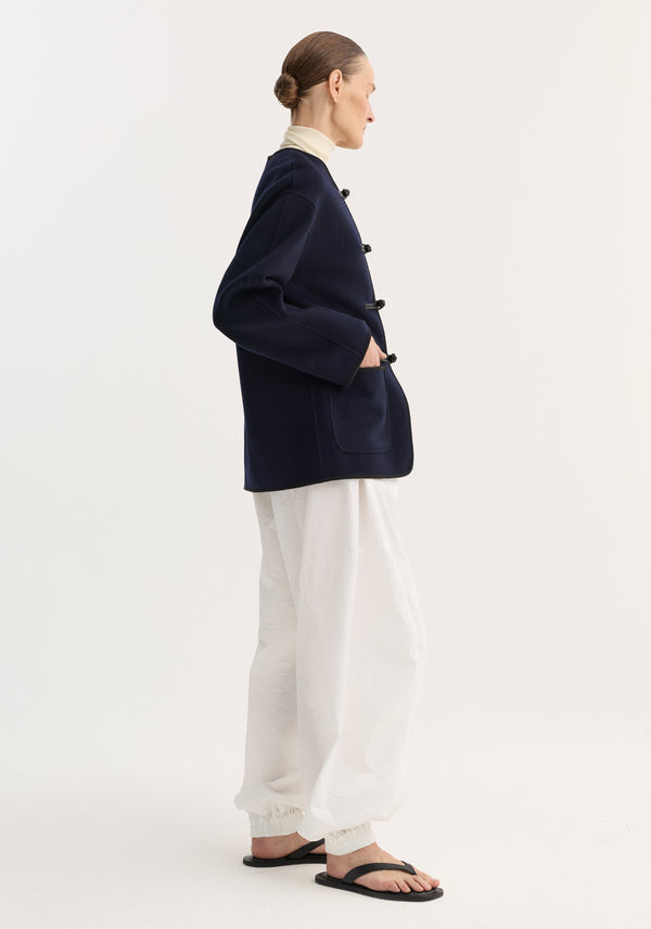 Side-profile of model wearing Mandarin double-faced wool jacket in navy, featuring pankou closure, embroidered edges, and patch pockets, styled with casual trousers and sandals.