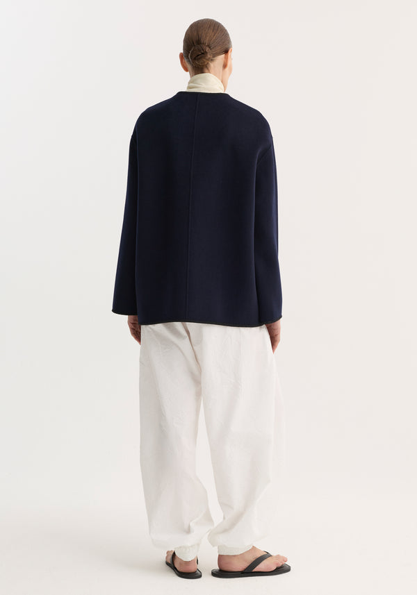 Back view of a model in the Mandarin double-faced wool jacket, showcasing its loose fit and embroidered edges, with a layered turtleneck and relaxed white pants.
