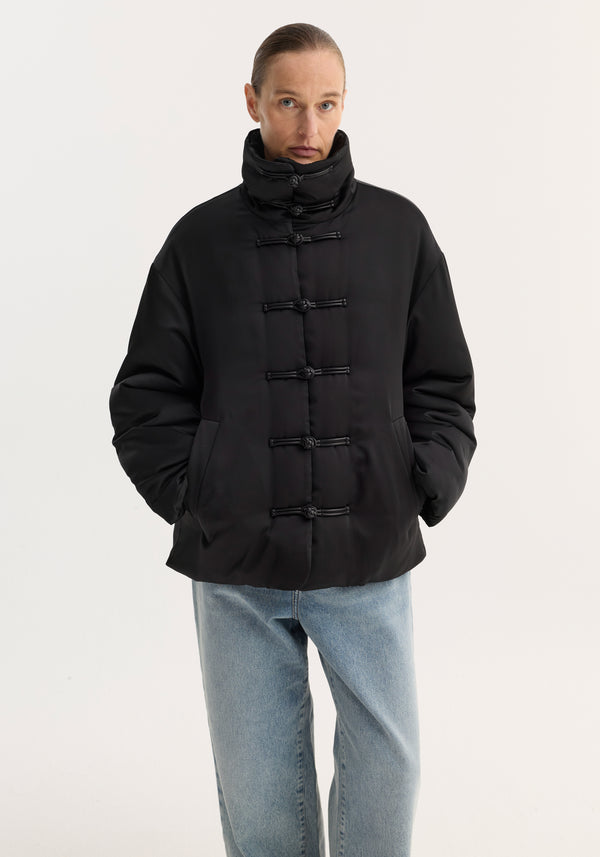 Person models the Padded puffer jacket | black, featuring a quilted design, high padded collar, toggle closures, and slanted pockets, paired with light-wash jeans.