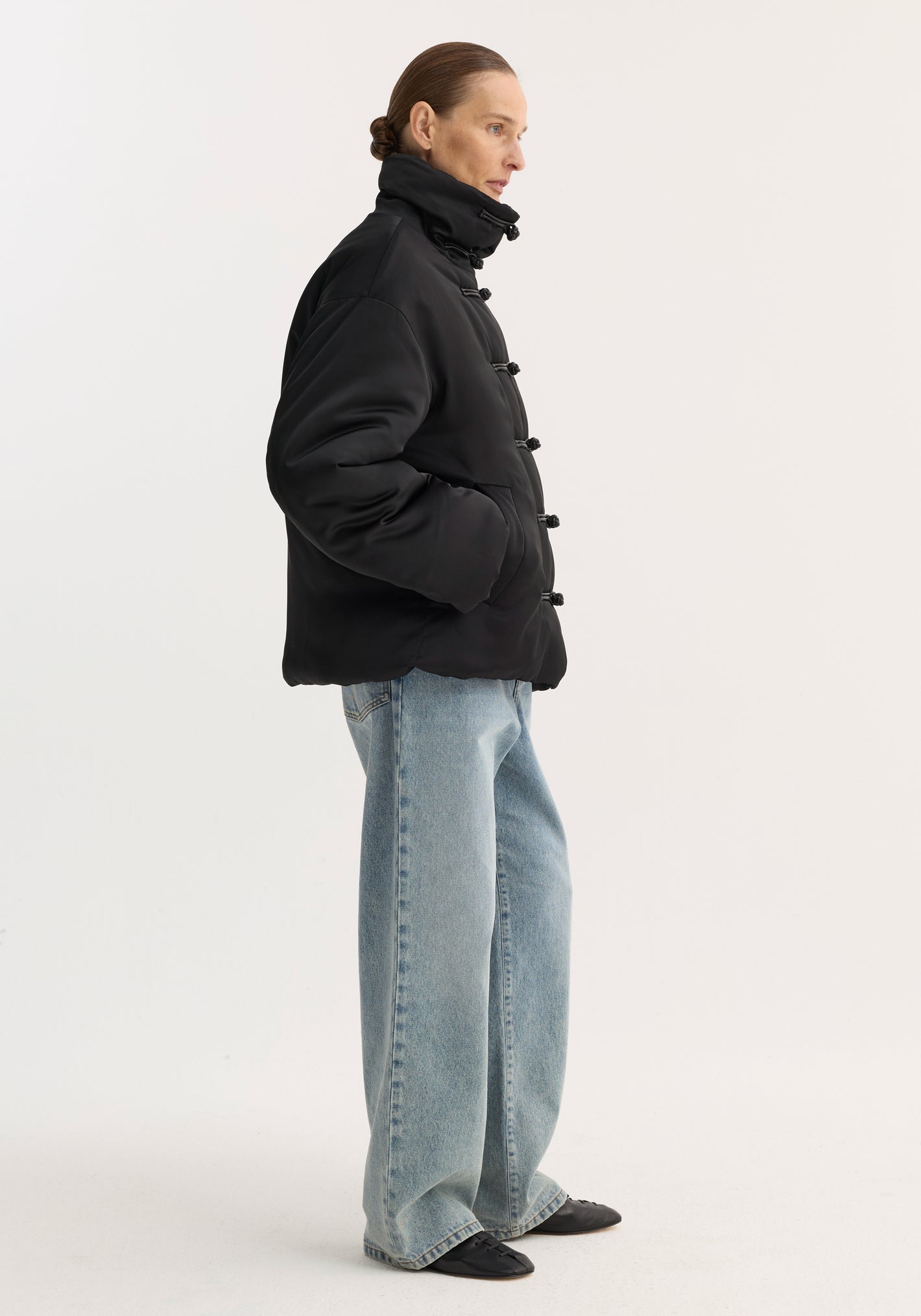 Person in profile wearing a padded puffer jacket | black with a high collar, pankou closures, and slanted pockets, paired with wide-leg jeans and low-profile shoes.
