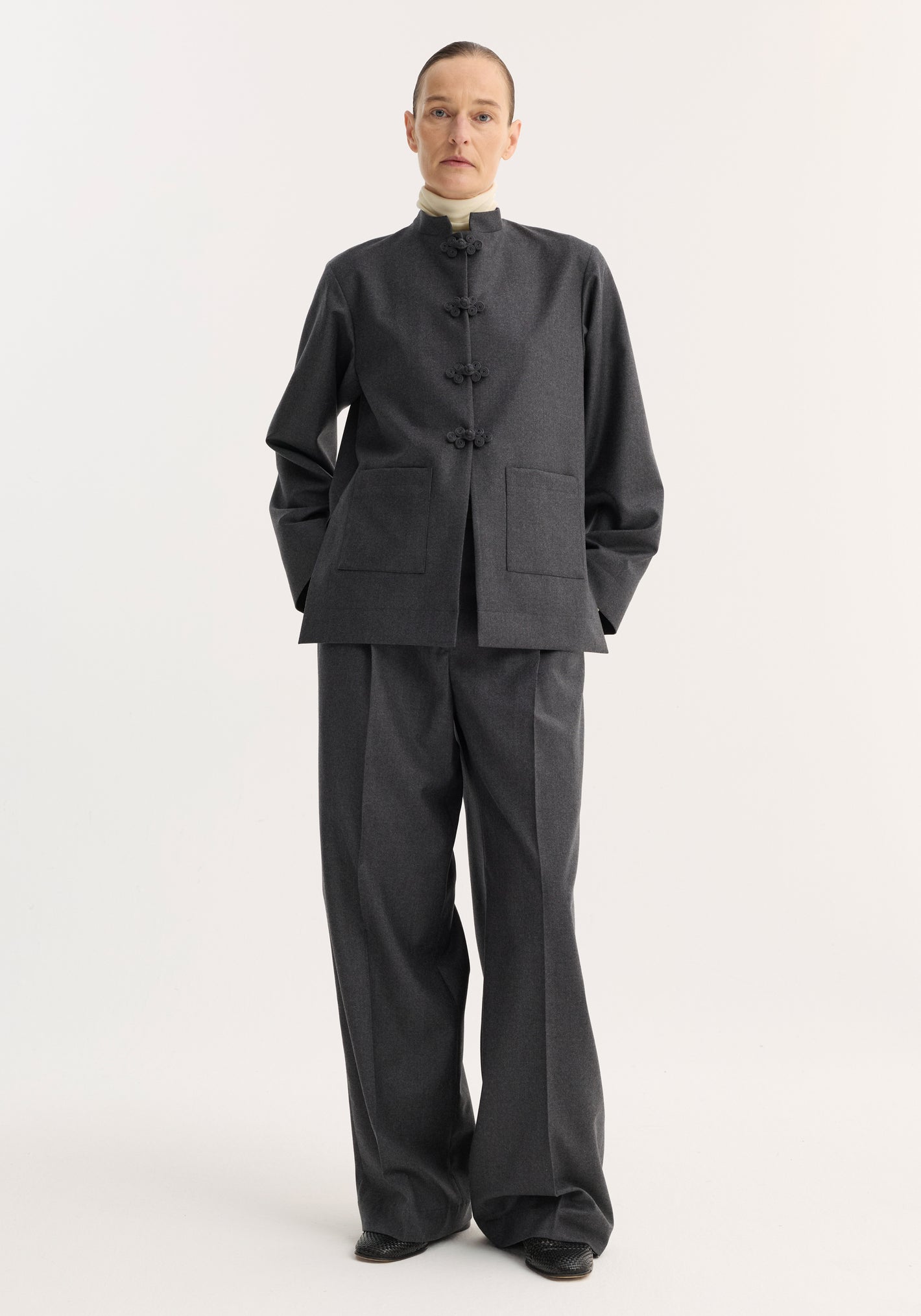 Model wearing the Mandarin jacket | dark grey melange, featuring a mandarin collar, pankou closure, and patch pockets, paired with matching trousers over black shoes.
